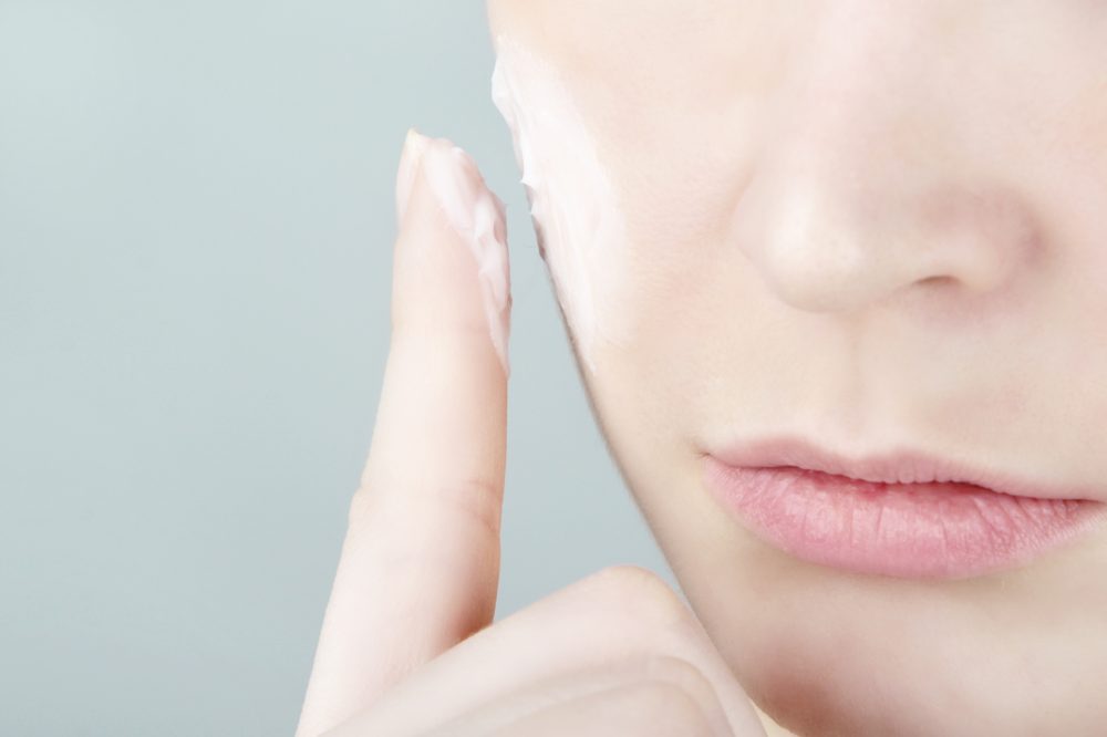 Young woman applies moisturizer cream onto her face.